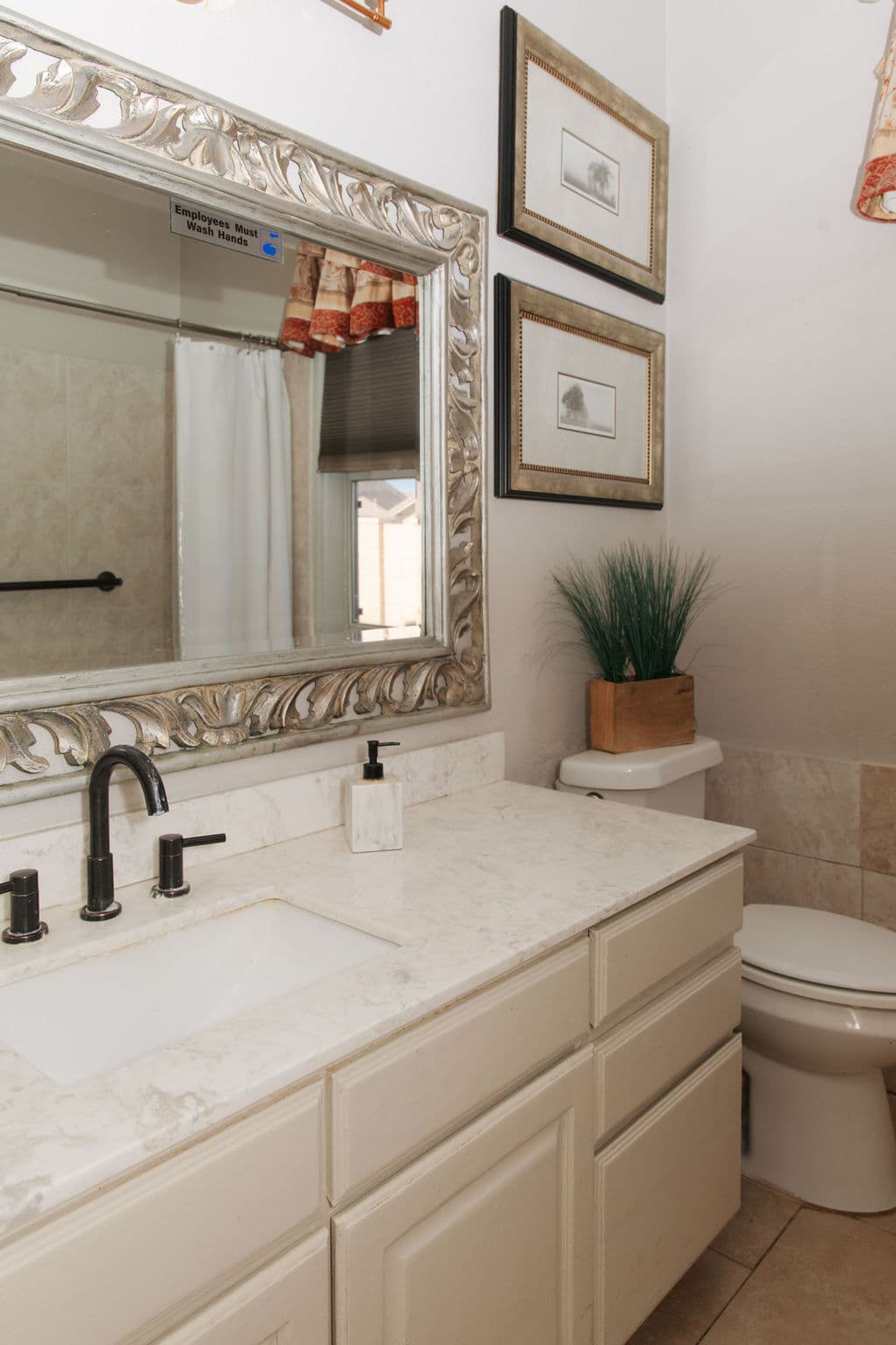 A bathroom with a marble-topped vanity, an ornate silver mirror, framed prints and a grab bar beside the toilet.