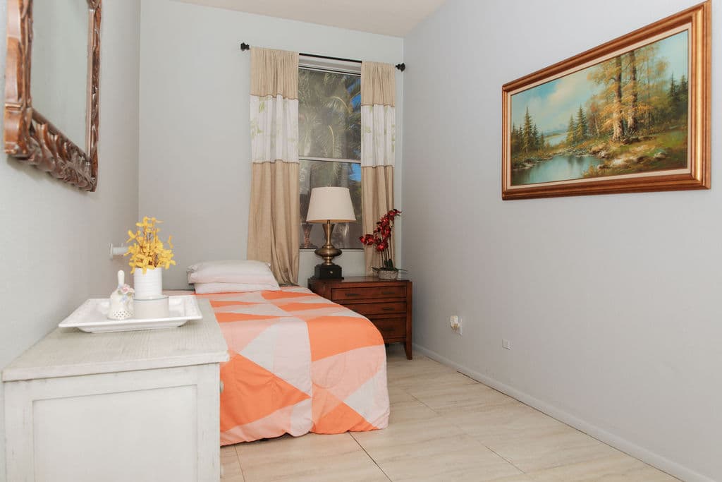 A bright bedroom with a peach-and-white quilt, a bedside lamp, a framed landscape painting and a window with garden views.