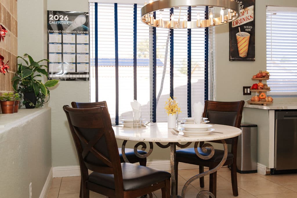A round marble breakfast table set with plates and folded napkins beside a sunny window, with a bowl of fruit on the counter.