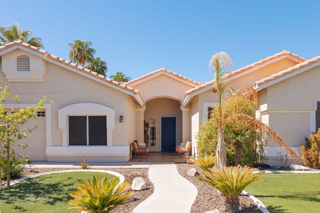 The front of the Liora home: a single-story stucco house with a terracotta tile roof, a blue front door, palm trees and a curved front walk.