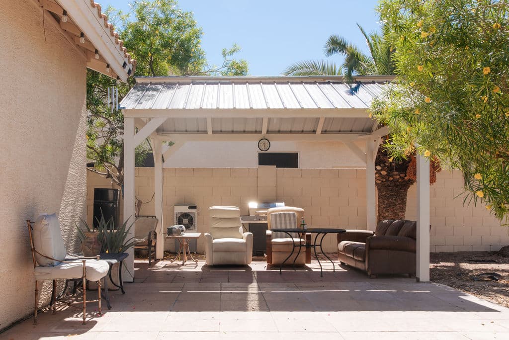 A shaded pergola in the backyard furnished with recliners, a sofa and a dining table, set among palms and trees.